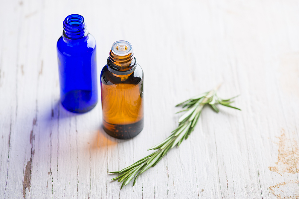 Amber and blue glass bottles containing natural fragrance oils with a fresh rosemary sprig on a wooden surface.