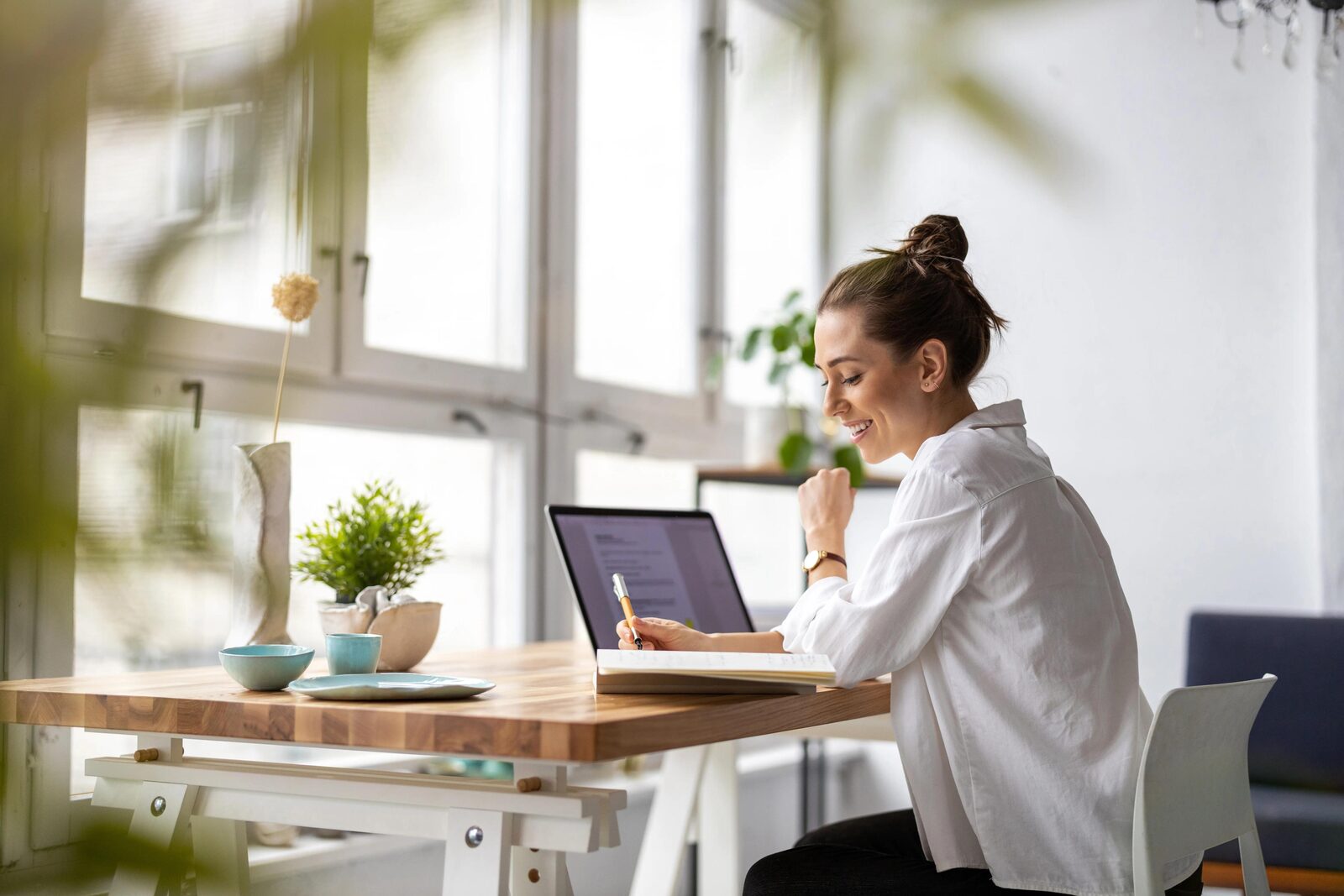 Woman writing in a notebook at a wooden desk positioned beside a bright window